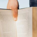 Opportunities - Crop unrecognizable male student in casual outfit standing with book in hand on green spring meadow with sunshine lighting up pages of novel