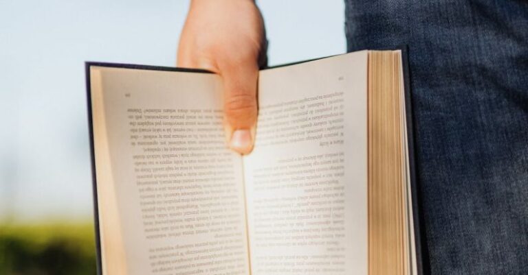 Opportunities - Crop unrecognizable male student in casual outfit standing with book in hand on green spring meadow with sunshine lighting up pages of novel